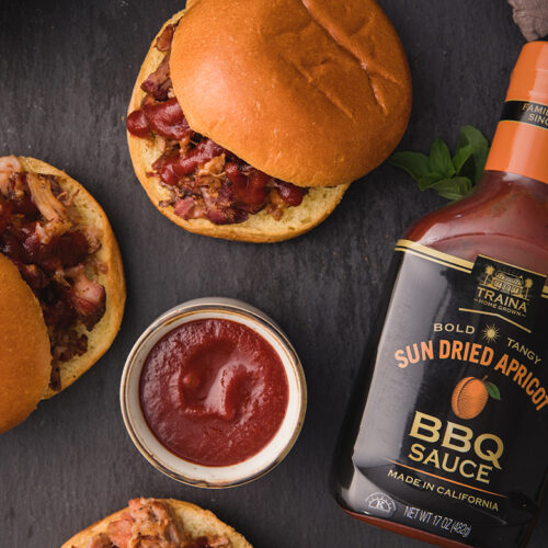 Overhead view of Sun Dried Apricot BBQ Sauce bottle next to pulled pork sandwiches.