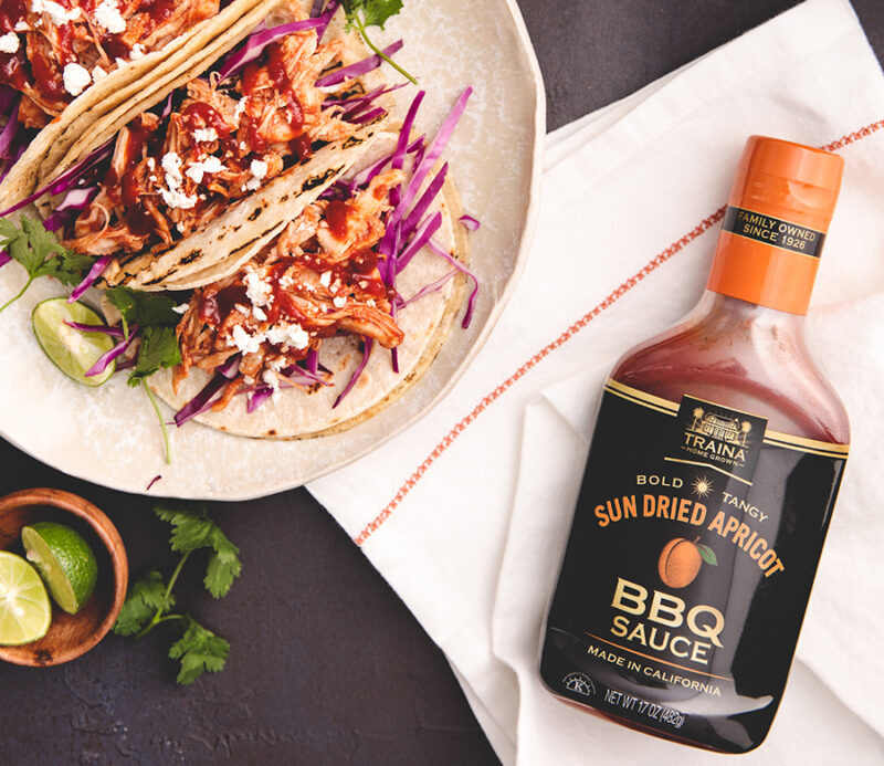 Overhead view of Sun Dried Apricot BBQ Sauce bottle beside marinated shredded chicken tacos.