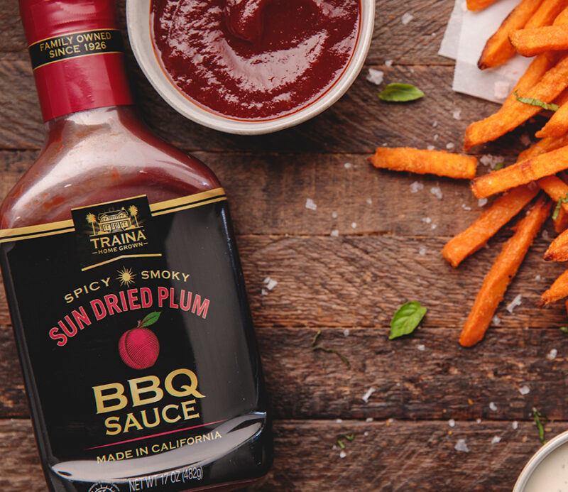 Overhead view of a textured brown wood board featuring a bottle of Sun Dried Plum BBQ Sauce, a small tub showing the sauce's thick texture, and scattered sweet potato fries, suggesting use as a delicious topping.